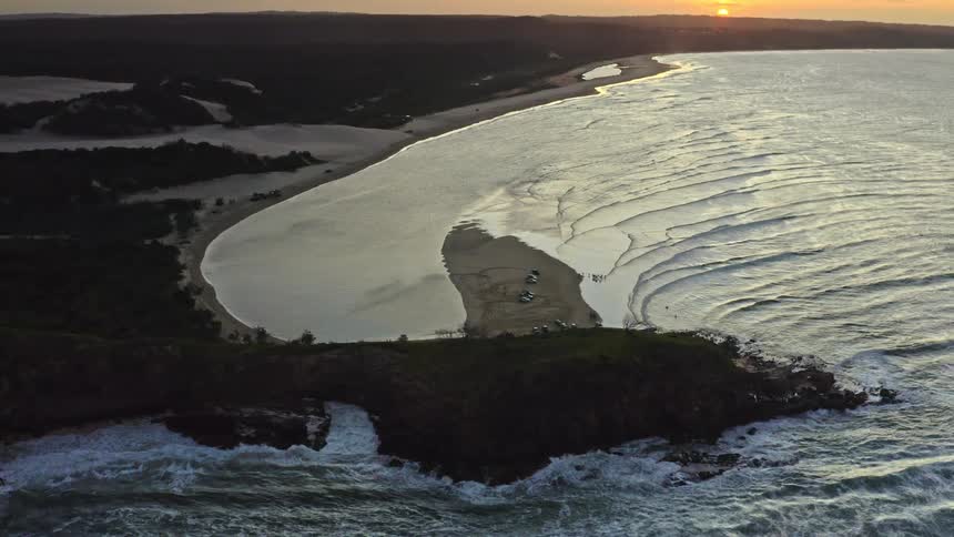 A static aerial view captures a dramatic coastline at sunset, with waves crashing on a rocky headland next to a vast sandy bay