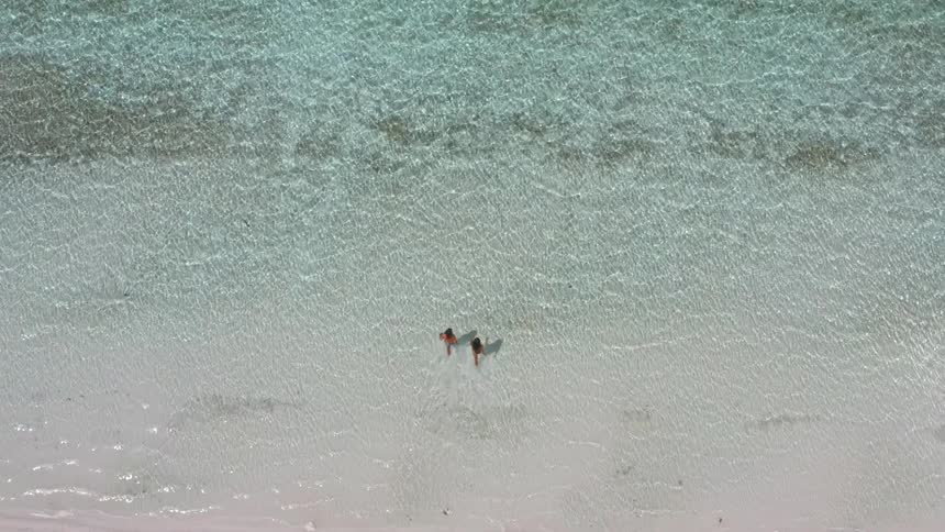 An aerial top-down shot shows a couple walking from the white sand beach into the crystal-clear turquoise ocean