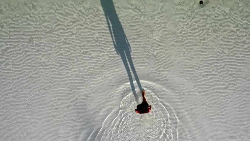An artistic top-down aerial view shows a woman's shadow preceding her as she walks through clear shallow water, creating mesmerizing ripples over the white sand