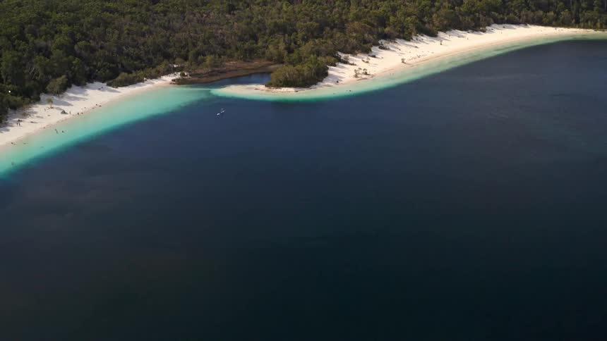 An incredible aerial reveal of a stunning tropical freshwater lake with deep blue water, a white sandy shore, and a lush forest