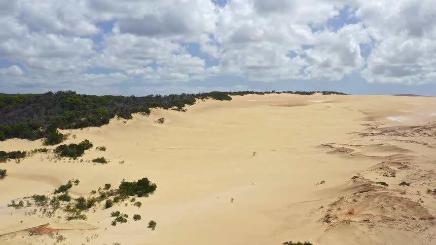 A vast desert of golden sand dunes stretches to the horizon as a drone slowly ascends, revealing tiny figures walking across the immense landscape