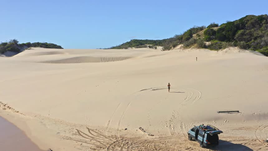 Aerial pullback shot of a woman walking across vast coastal sand dunes as off-road vehicles are parked on the beach