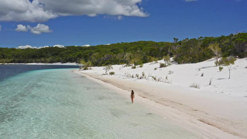 Drone follows a woman in a bikini walking along a breathtaking white sand beach next to crystal clear turquoise water on a sunny day