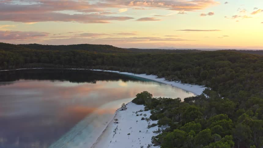 A tranquil aerial pullback reveals a stunning freshwater lake with a white sandy shoreline nestled in a dense forest at sunset