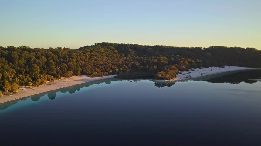 A stunning aerial pullback reveals a tranquil, crystal-clear lake reflecting the dense forest and white sand beaches during a golden sunrise