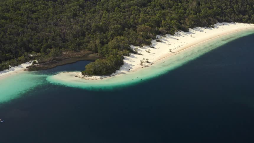 An aerial view tilts up from a deep blue lake to reveal a stunning white sand beach surrounded by a dense, green forest