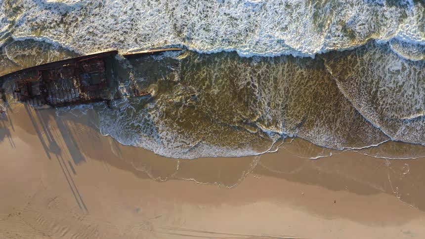 Aerial top-down pull-out reveals a historic shipwreck on a sandy beach with ocean waves crashing against its rusty hull at sunrise