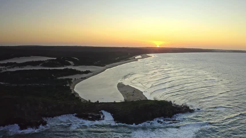 Majestic aerial pullback reveals a stunning coastal landscape with a wide sandy inlet and rolling ocean waves at golden hour sunset
