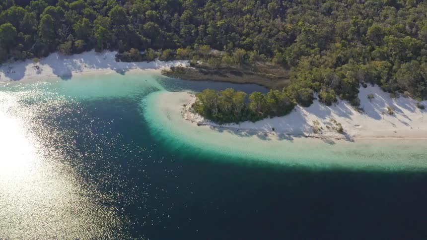 Rising aerial shot reveals the spectacular natural beauty of a turquoise freshwater lake with white sand shores surrounded by a dense forest