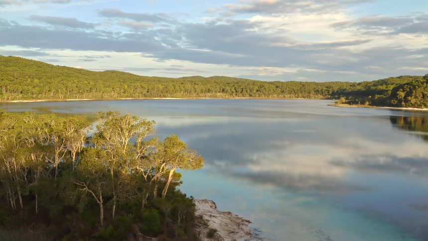A serene aerial ascent reveals a vast, tranquil lake at golden hour, its calm surface perfectly mirroring the sky and lush surrounding forest
