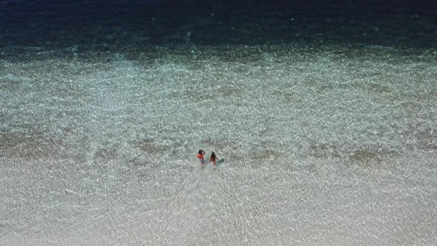 An aerial top-down view of two women wading from a white sandy beach into calm, crystal-clear turquoise ocean waters