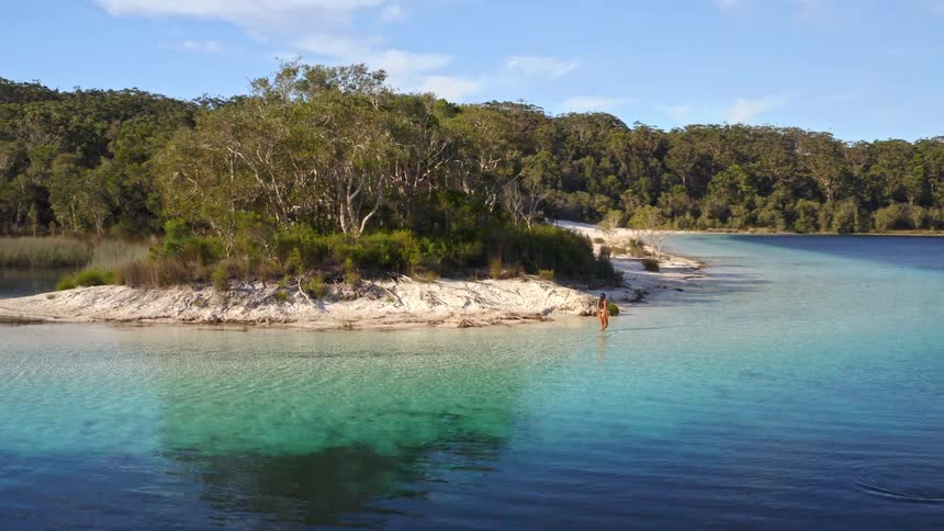 A woman wades in the crystal clear turquoise water of a remote tropical lake surrounded by a lush forest and white sand