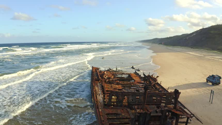 An expansive aerial shot pulls back along a beautiful coastline to reveal a large, rusty shipwreck being lapped by ocean waves