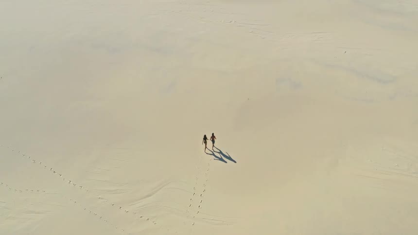 An aerial high-angle view follows a young couple in swimwear as they walk across a vast, empty sand dune, leaving a trail of footprints behind