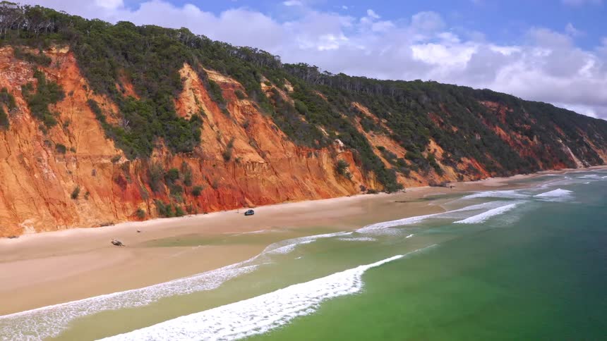 A breathtaking aerial shot reveals a 4x4 vehicle parked on a remote sandy beach beside vibrant, colorful cliffs and the tranquil ocean