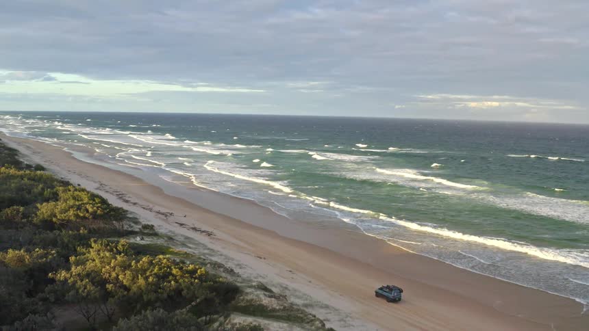 An aerial view captures a 4x4 vehicle driving along a vast, sandy coastline as waves crash on the shore during a beautiful sunrise