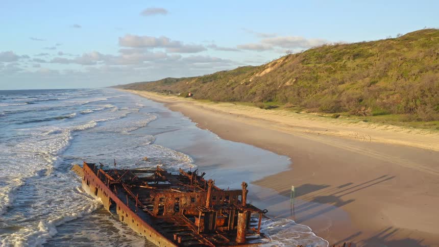 An aerial pullback reveals the rusted Maheno shipwreck beached on the sandy shores of Fraser Island as gentle waves wash over it at sunrise