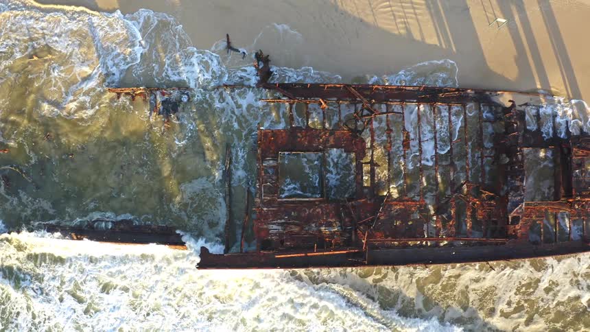 A drone provides a top-down view as it slowly pans across the rusted skeleton of a shipwreck, beached on the sand as waves crash through its decaying frame