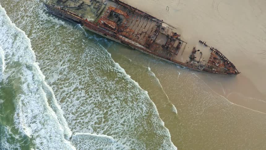An aerial top-down shot of the iconic Maheno shipwreck on Fraser Island slowly pulls back, revealing the vast, beautiful coastline as waves crash against the rusted hull