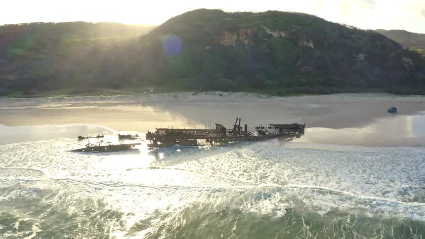 An aerial perspective reveals a historic shipwreck resting on a remote beach, with gentle ocean waves lapping at its rusted hull during a beautiful golden hour