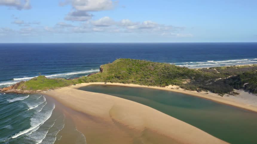A slow aerial pullback reveals a stunning coastal landscape where a sandy spit separates a calm lagoon from the deep blue ocean