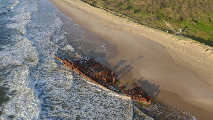 A stunning aerial view tilts up to reveal the iconic Maheno Shipwreck beached on the sandy shores of Fraser Island, Australia, as waves crash against its rusty hull