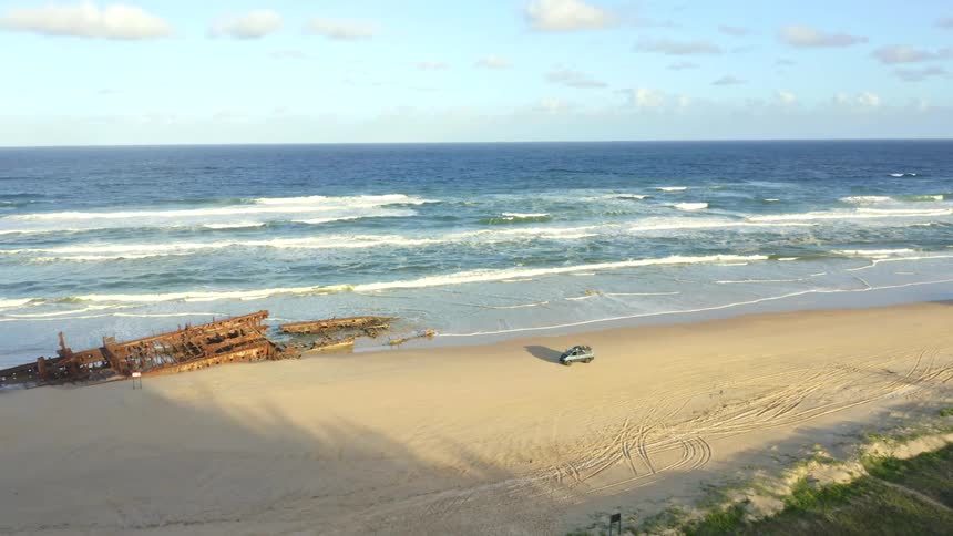 Slow aerial pan across a beautiful sandy beach reveals a large, rusted shipwreck with off-road vehicles parked on the shore