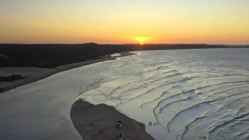 Epic aerial view of a wide river mouth meeting the ocean waves along a sandy coastline during a beautiful golden sunset
