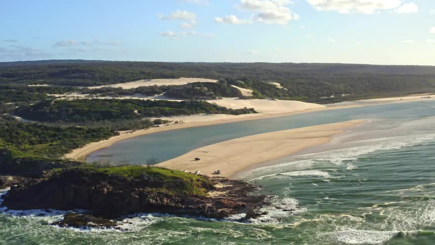 An incredible aerial view reveals a stunning coastal landscape where a rocky headland meets a tranquil estuary, sandy beach, and vast dunes