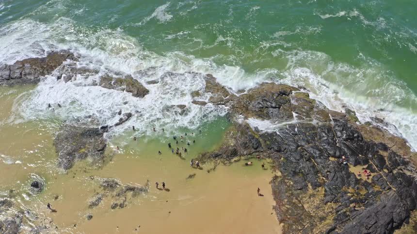 An ascending top-down aerial view shows people swimming in a tidal pool as ocean waves crash along the rocky shore