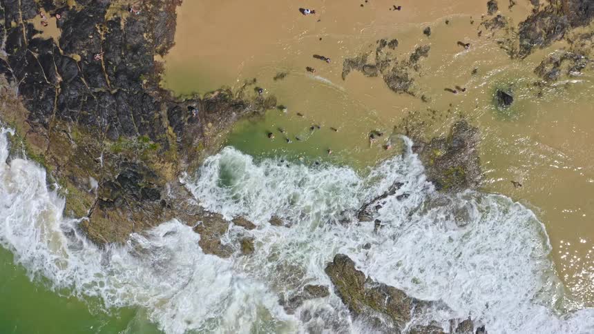 An overhead aerial view captures people enjoying a calm rock pool as a powerful ocean wave crashes over the rocks and into the water