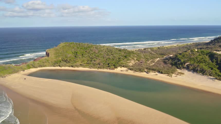 Slowly pulling back aerial shot over a beautiful estuary and sand dunes that lead out to the vast blue ocean on a sunny day
