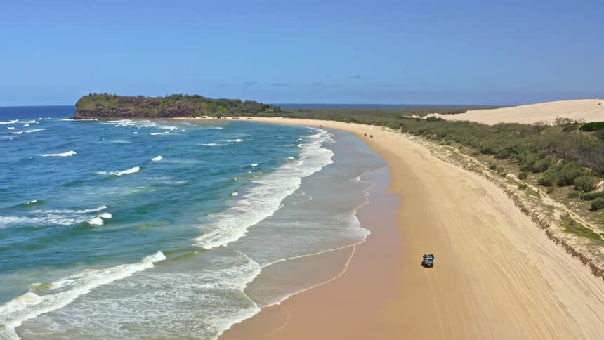 A cinematic aerial shot follows a 4x4 vehicle driving along a vast, scenic beach with golden sand and turquoise ocean waves under a clear blue sky