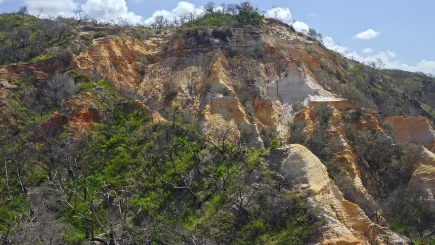 A slow pullback shot reveals the stunning, multi-colored sand cliffs and surrounding lush, green bushland under a partly cloudy sky