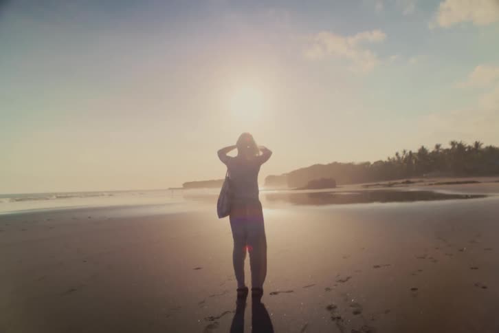 A silhouetted woman stands on a vast, dark sand beach, enjoying the golden sunset over the ocean as the camera slowly pulls back