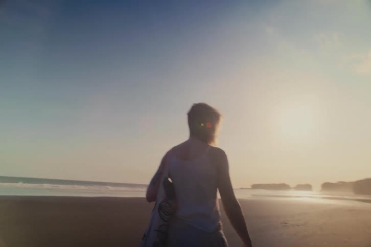 Slow motion shot from behind of a woman with a tote bag walking along a tranquil sandy beach during a beautiful golden sunset