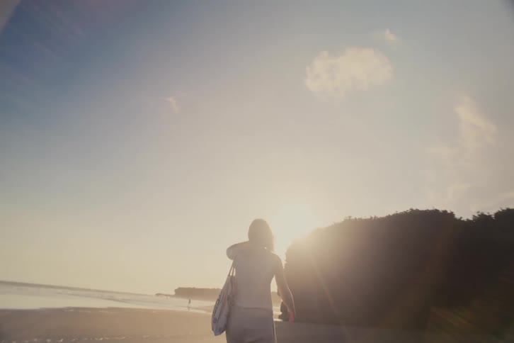 A woman walks in slow motion along a sandy beach as the golden sun sets over the ocean