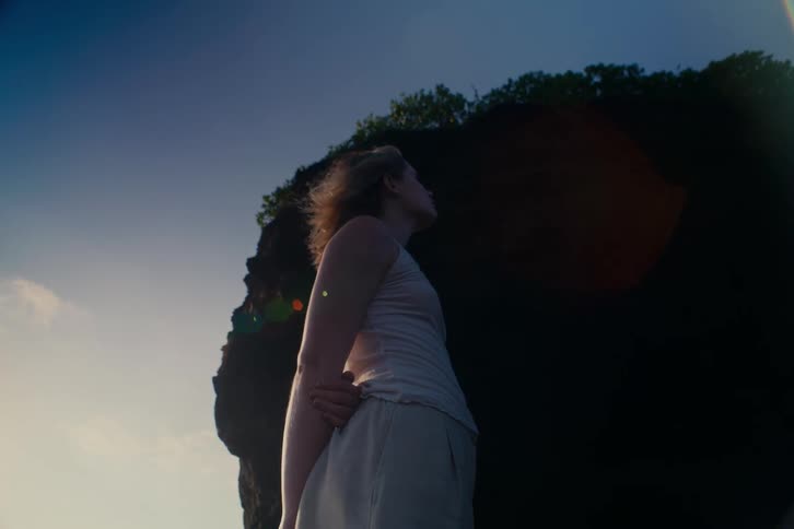 A pensive young woman stands against a silhouetted rock formation as the wind blows through her hair in slow motion