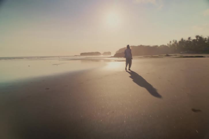 A man takes a peaceful and solitary walk along a vast, reflective tropical beach during a beautiful golden hour