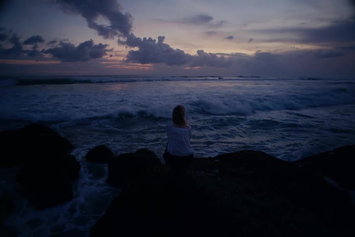A woman sits alone on a rock, watching the ocean waves crash against the shore during a beautiful twilight