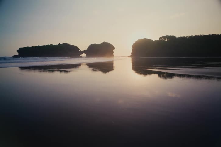 A serene sunset over a reflective wet sand beach with silhouetted rock formations on the horizon
