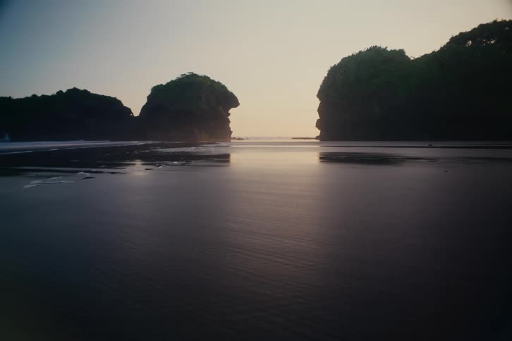 A tranquil sunset reflects on the wet, dark sand of a secluded beach cove flanked by rocky islands