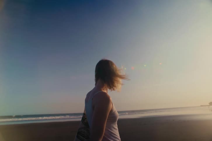 A slow-motion, over-the-shoulder shot captures a woman with windblown hair standing on a beach at sunset, gazing thoughtfully at the ocean