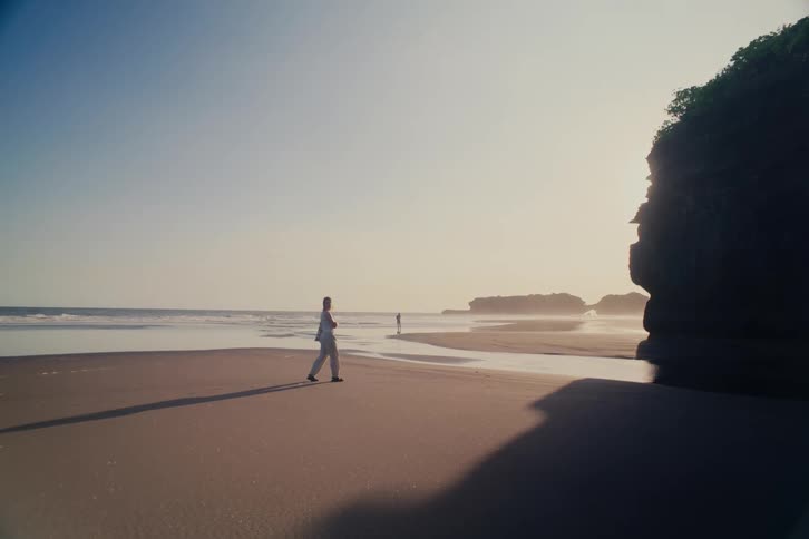 A solitary person walks along a vast, tranquil beach at sunrise, stopping to look out at the ocean next to a large cliff