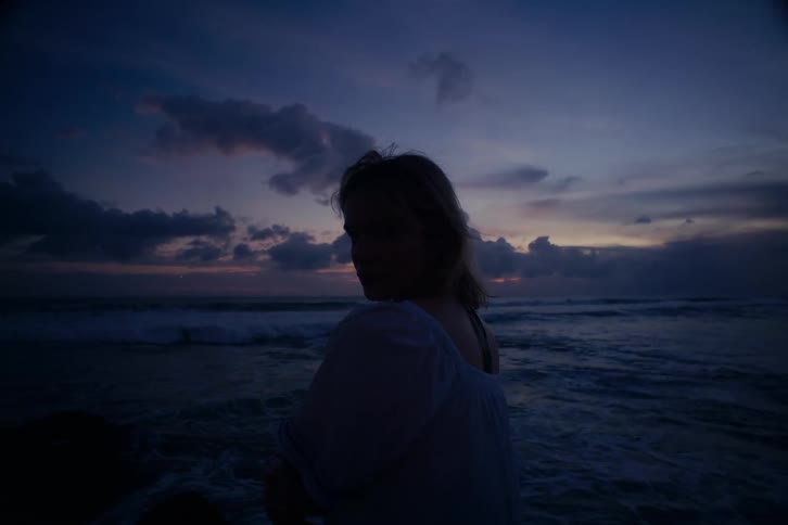 A silhouetted woman turns to look over her shoulder while standing on the beach at twilight