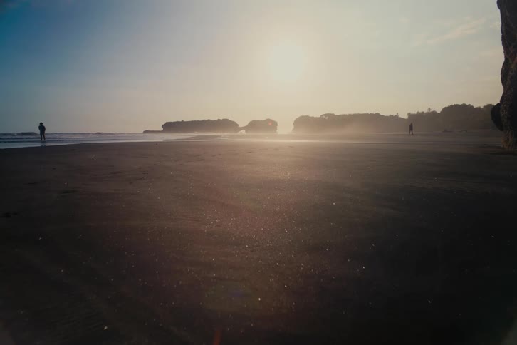 A serene, hazy golden hour blankets a vast dark sand beach with distant silhouetted figures and cliffs