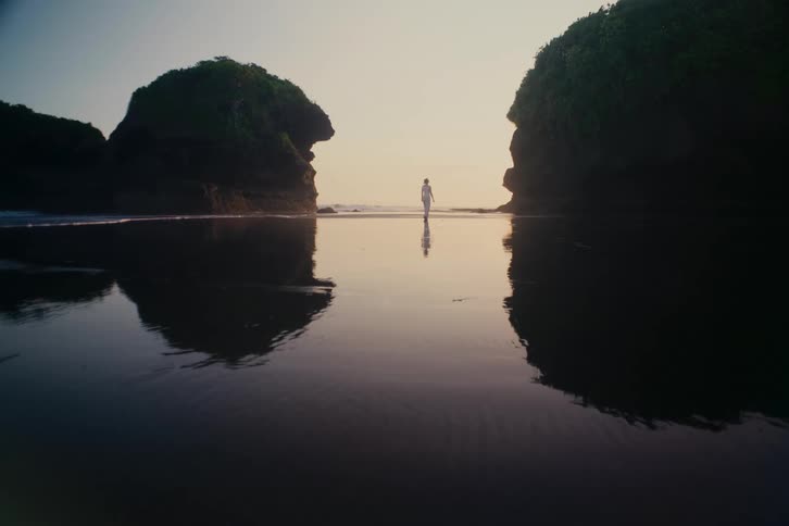 A lone figure stands on a secluded, reflective beach between two silhouetted rock formations at sunset