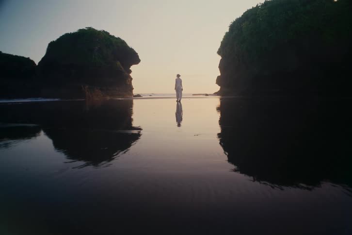 A solitary figure in white is reflected in the wet sand as they walk on a serene beach between two large rock formations at dawn
