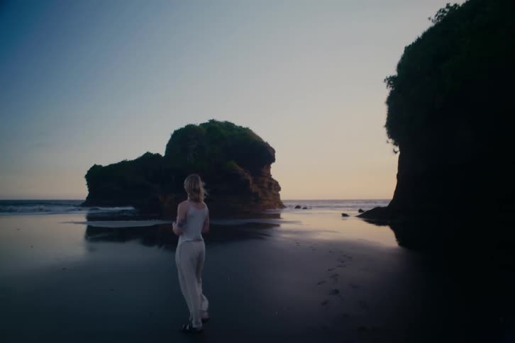 A young woman walks in slow motion along a serene, dark sand beach with large rock formations at dusk