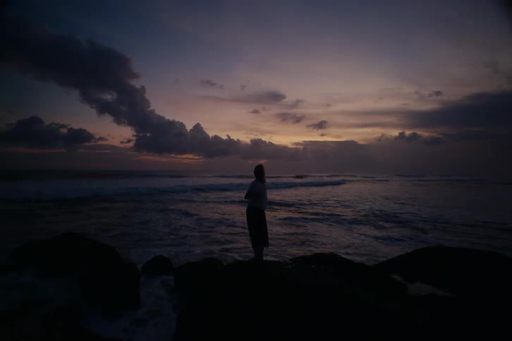 Silhouette of a woman standing on rocks by the ocean, peacefully watching the waves under a dramatic twilight sky